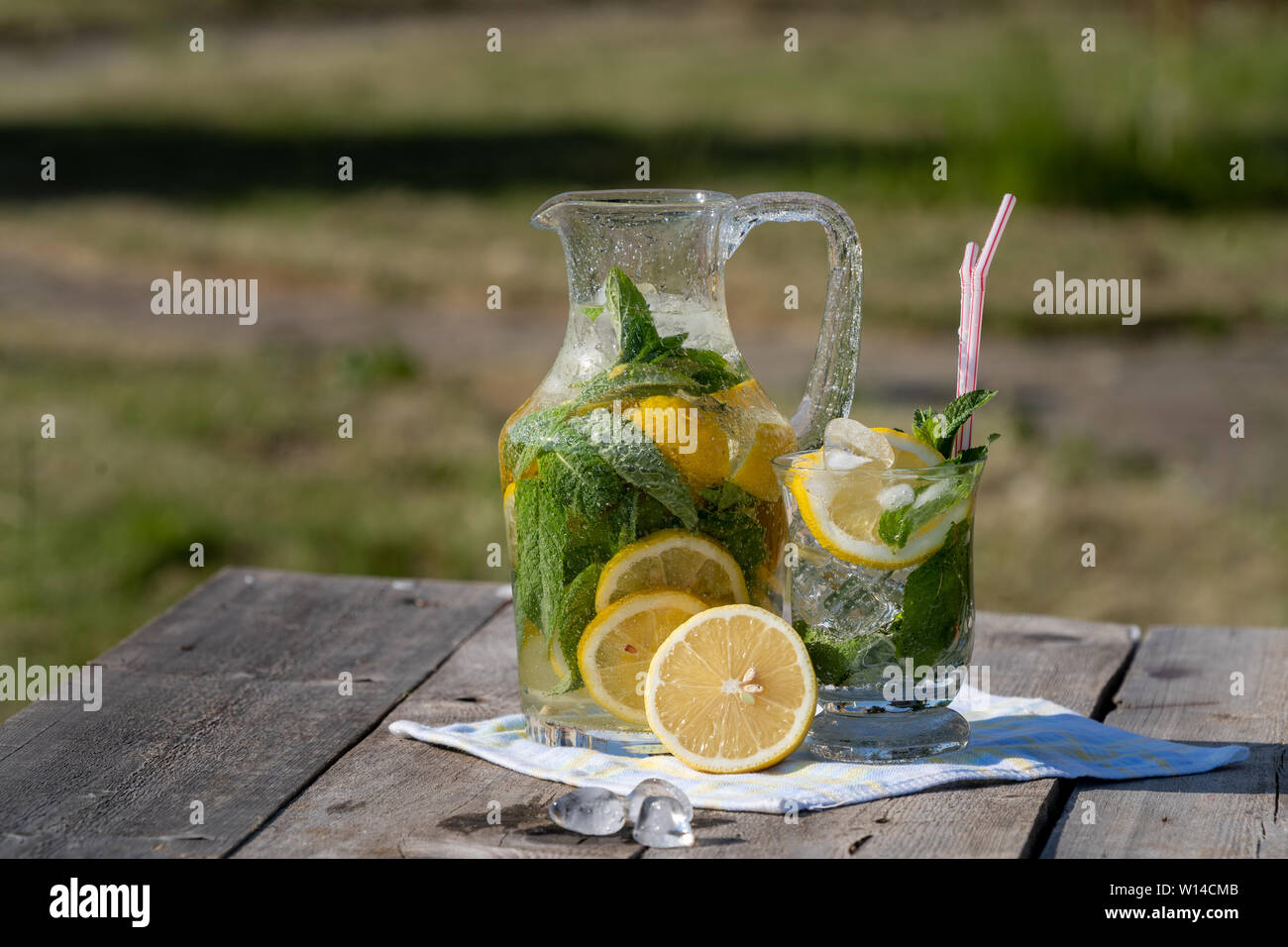 Concetto di estate. Limonata fatta in casa con il limone, menta e ghiaccio in una caraffa e un vaso, su un vecchio tavolo di legno, all'aperto, sfocato sfondo verde. Foto Stock