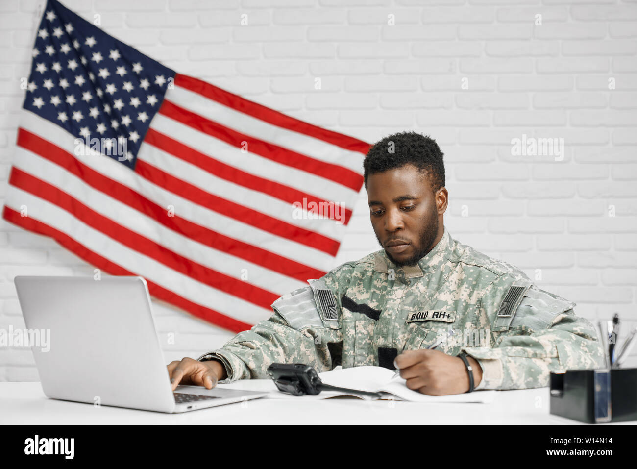 Soldato maschio o lavoratore di esercito americano seduta a tavola e la scrittura di documenti utilizzando laptop. American ranker seriamente guardando e sottoscrizione contra Foto Stock