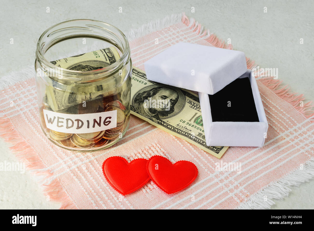 Parola di nozze, monete e il dollaro in un vasetto di vetro vicino a due cuori di seta e vuoto bianco anello di nozze box su una rosa orlata igienico. Risparmio di denaro per noi Foto Stock