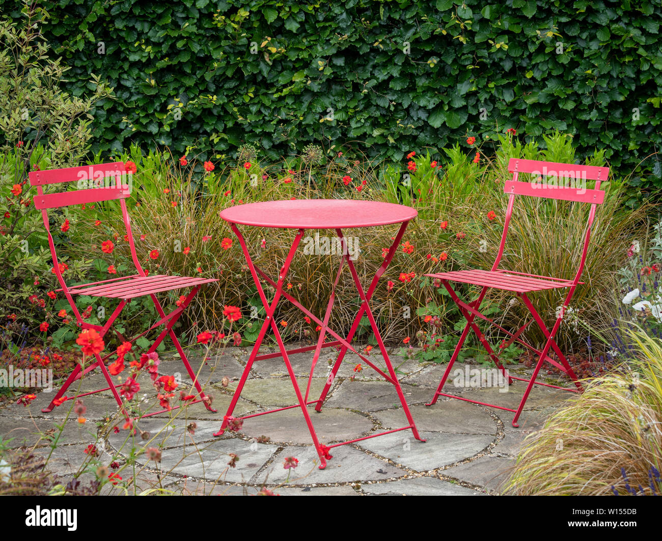 Zona appartata del giardino con un piccolo patio e due sedie rosse con tavolo in metallo circondato da fiori estivi e graminacee. Foto Stock