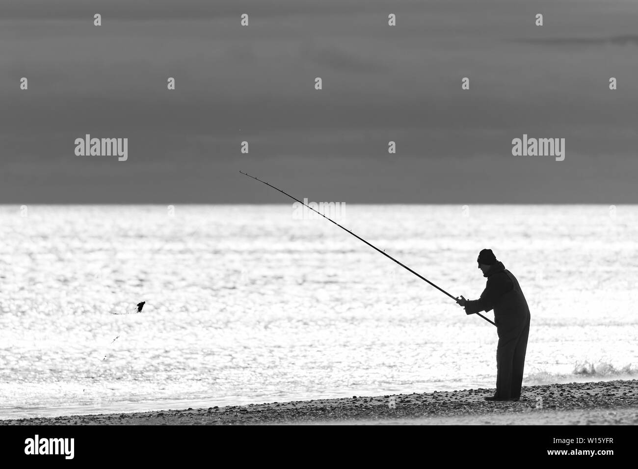 Immagine in bianco e nero di spiaggia pescatore con asta sollevata la preparazione di gettare la sua esca fuori ad un mare calmo Foto Stock