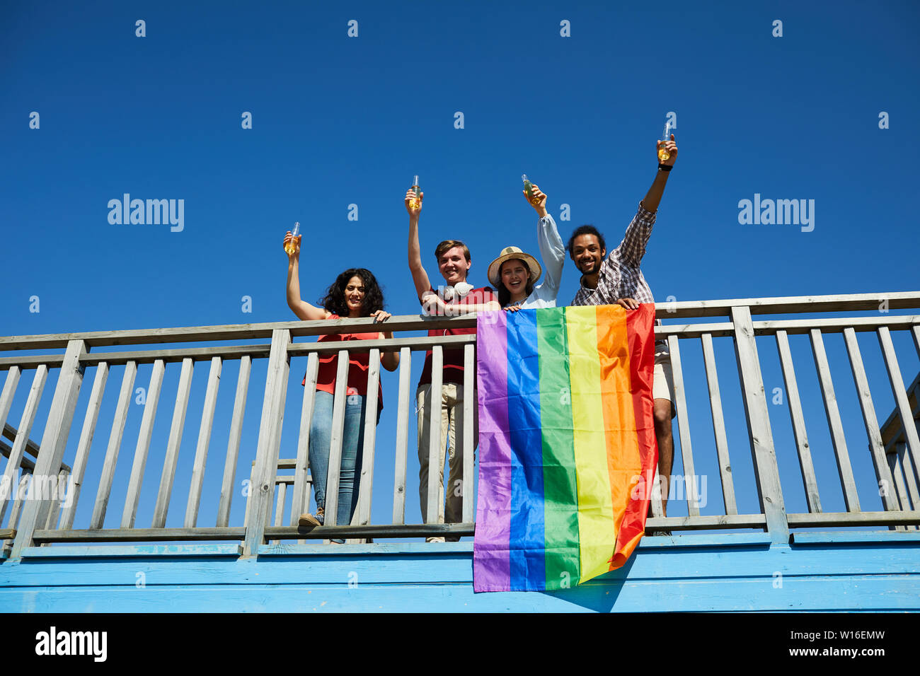 Angolo basso ritratto di felice giovani in piedi sul ponte e il tifo con bandiera arcobaleno in primo piano, spazio di copia Foto Stock