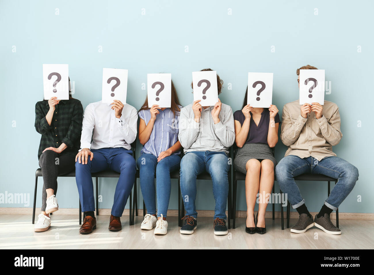 Giovani volti nascosti dietro i fogli di carta con punti interrogativi in attesa per il colloquio di lavoro in interni Foto Stock