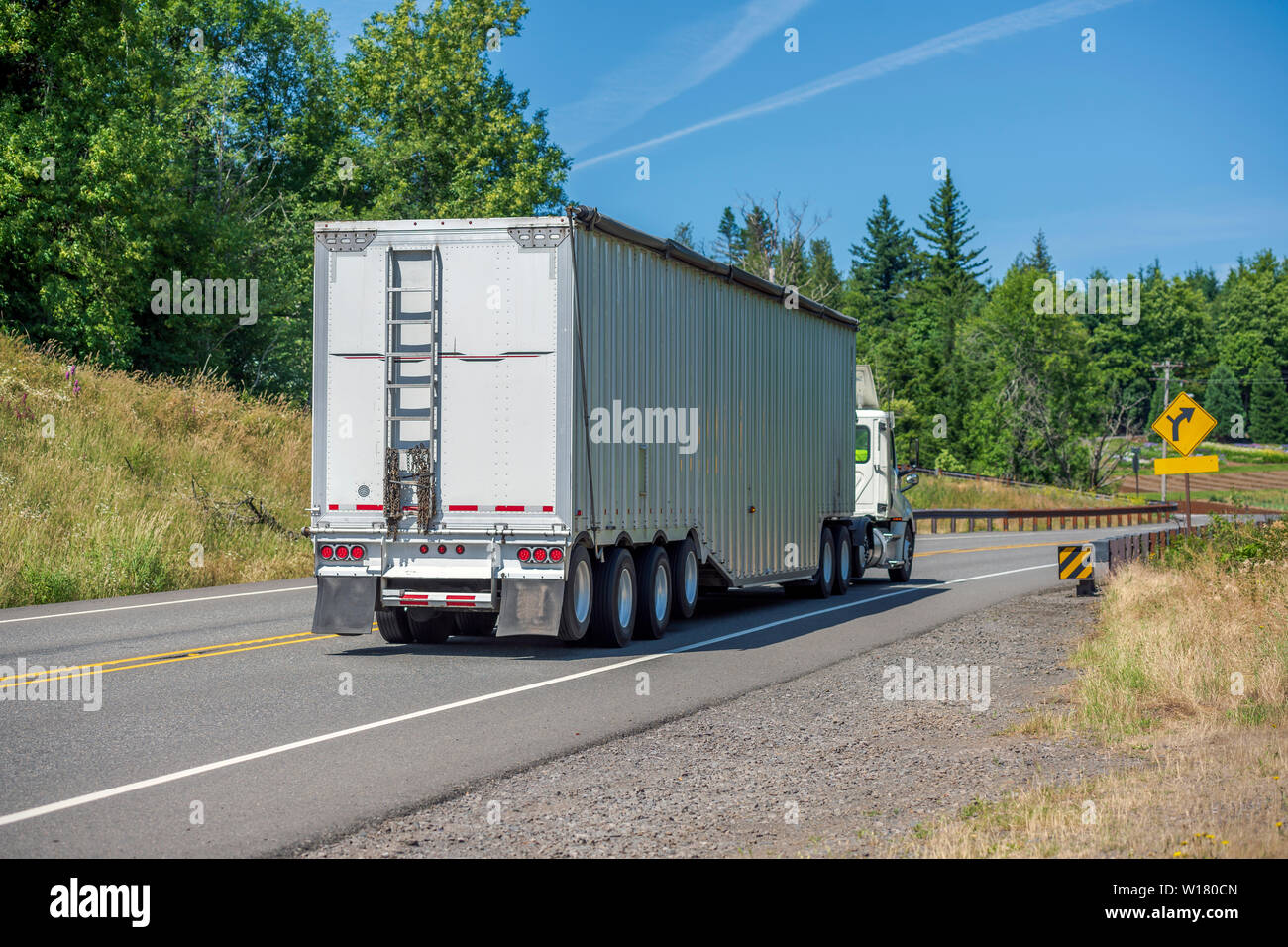 Big Rig bianco cabina giorno semi carrello con spoiler del tetto trasportano enormi coperte rinfusa semi rimorchio con scaletta sulla parete di fondo mobile sul vento di tornitura Foto Stock