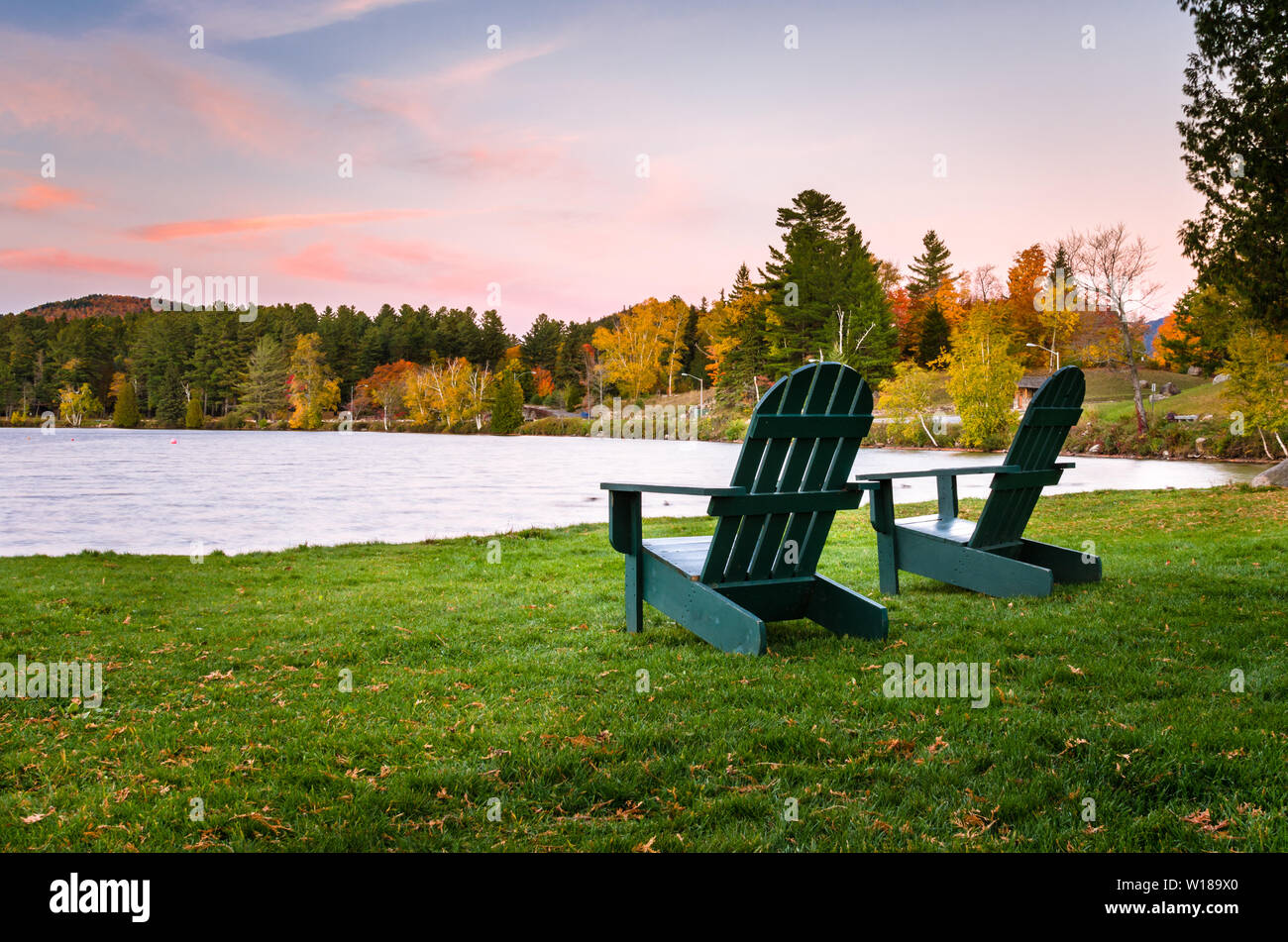 Due vuoti sedie Adirondack su un prato di fronte ad un lago al tramonto. Bellissimi colori autunnali. Foto Stock