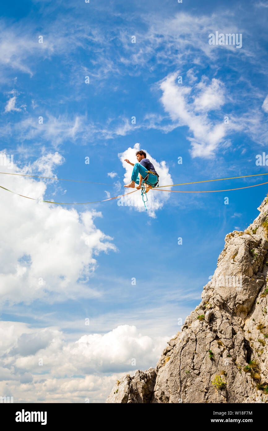 Giovane uomo su un highline sopra sharp scogliere frastagliate. Slackline in montagna con splendidi e verdi pendii e soffici nuvole in background Foto Stock