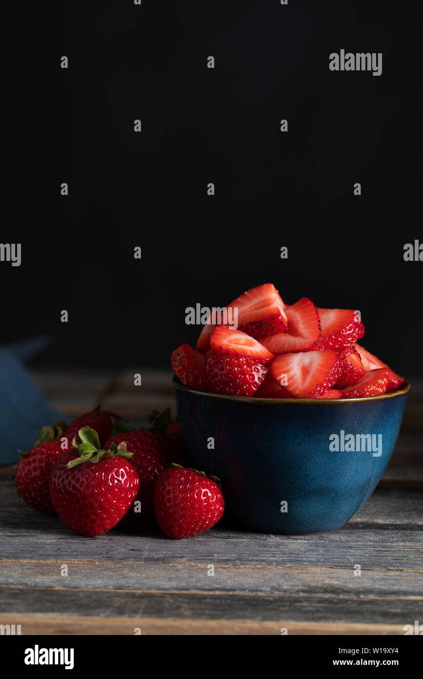 Fresche fragole a fette nella ciotola con le fragole intere accanto a loro, chiave di bassa illuminazione e orientamento verticale. Foto Stock