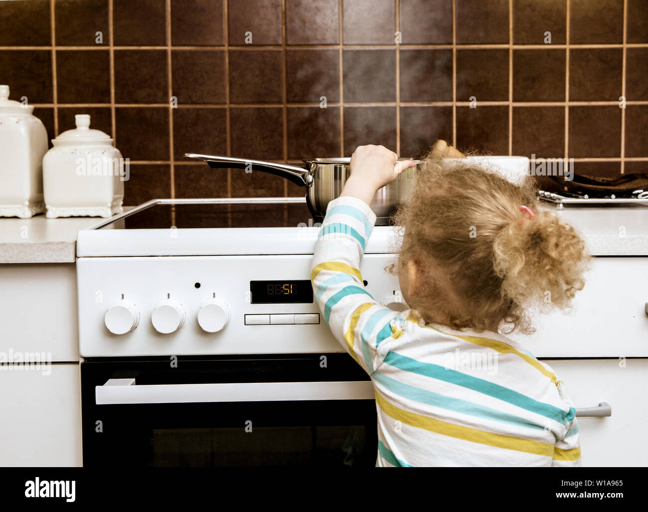 Incidente in attesa di accadere. Il Toddler girl tirando boiling pot di acqua su in cucina domestica. Luci a casa, ustioni, concetto( impostare, piatto è vuoto). Foto Stock