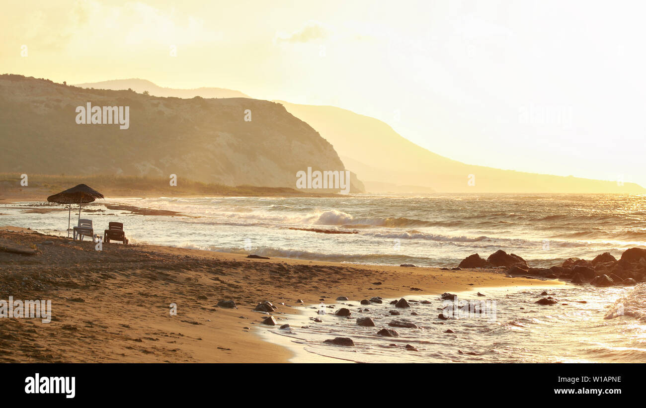 Due lettini sul vuoto spiaggia selvaggia accesa dal tramonto dorato. Piccole onde sulla roccia, colline in sfondo nebuloso Foto Stock