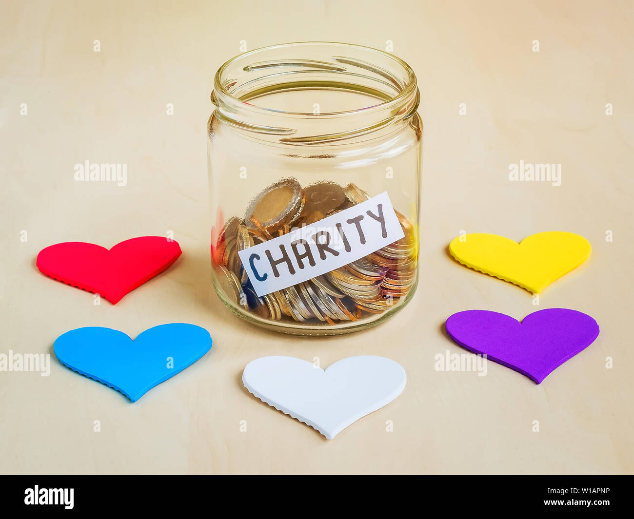 Un sacco di monete e di carità parola in un vasetto di vetro vicino a cinque cuori colorati. Il risparmio e la raccolta di fondi per beneficenza concetto. Foto Stock