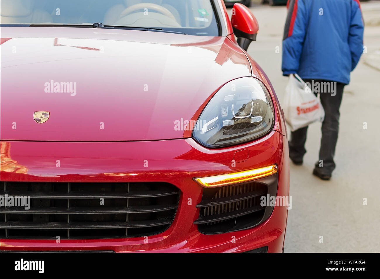 Varna, Bulgaria, 28 febbraio 2019. Un poco vestite senior uomo cammina per la strada passato un rosso Porsche auto. Concetto di lusso e di povertà. Il contrasto Foto Stock