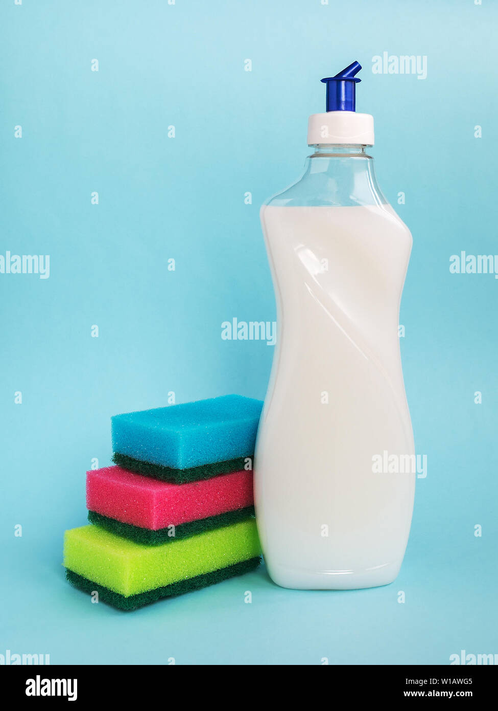 Flacone bianco con detersivo liquido per stoviglie e tre spugne di schiuma di diversi colori su sfondo blu. Prodotti chimici per cucina.detergente domestico. Foto Stock
