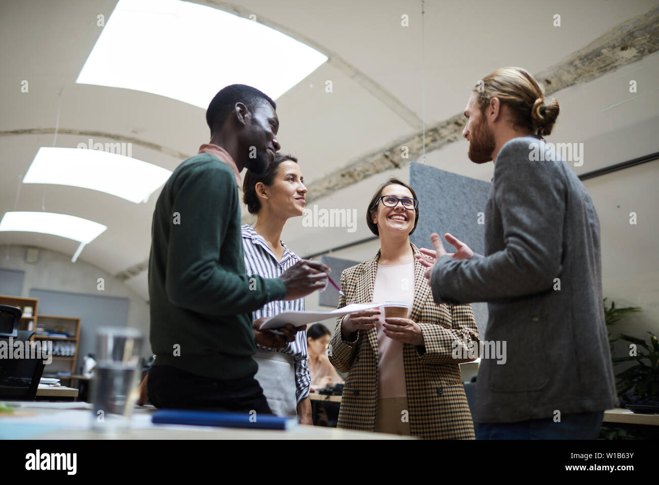 Basso angolo di visione a multi - gruppo etnico di uomini di affari che ridevano allegramente chiacchierando durante la pausa caffè in ufficio, spazio di copia Foto Stock