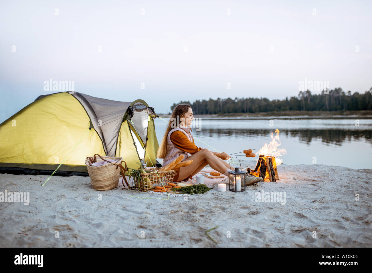 Giovane donna seduta davanti al camino, cucina salsicce, avente un picnic presso il campeggio sulla spiaggia la sera Foto Stock