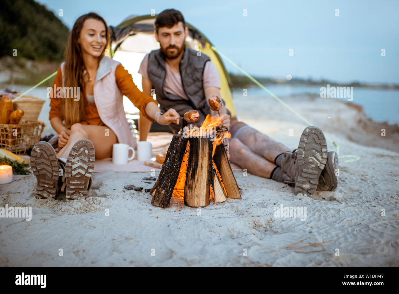 Giovane e allegro giovane la cottura di salsicce al caminetto, avente un picnic presso il campeggio sulla spiaggia la sera Foto Stock