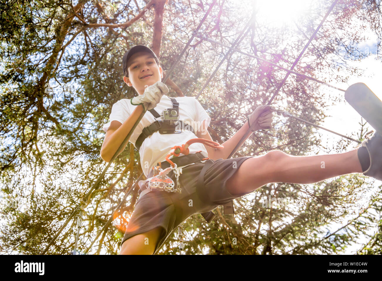 Adolescente con azione telecamera avente divertimento sulle corde alte corso, parco avventura arrampicata alberi nella foresta in estate Foto Stock