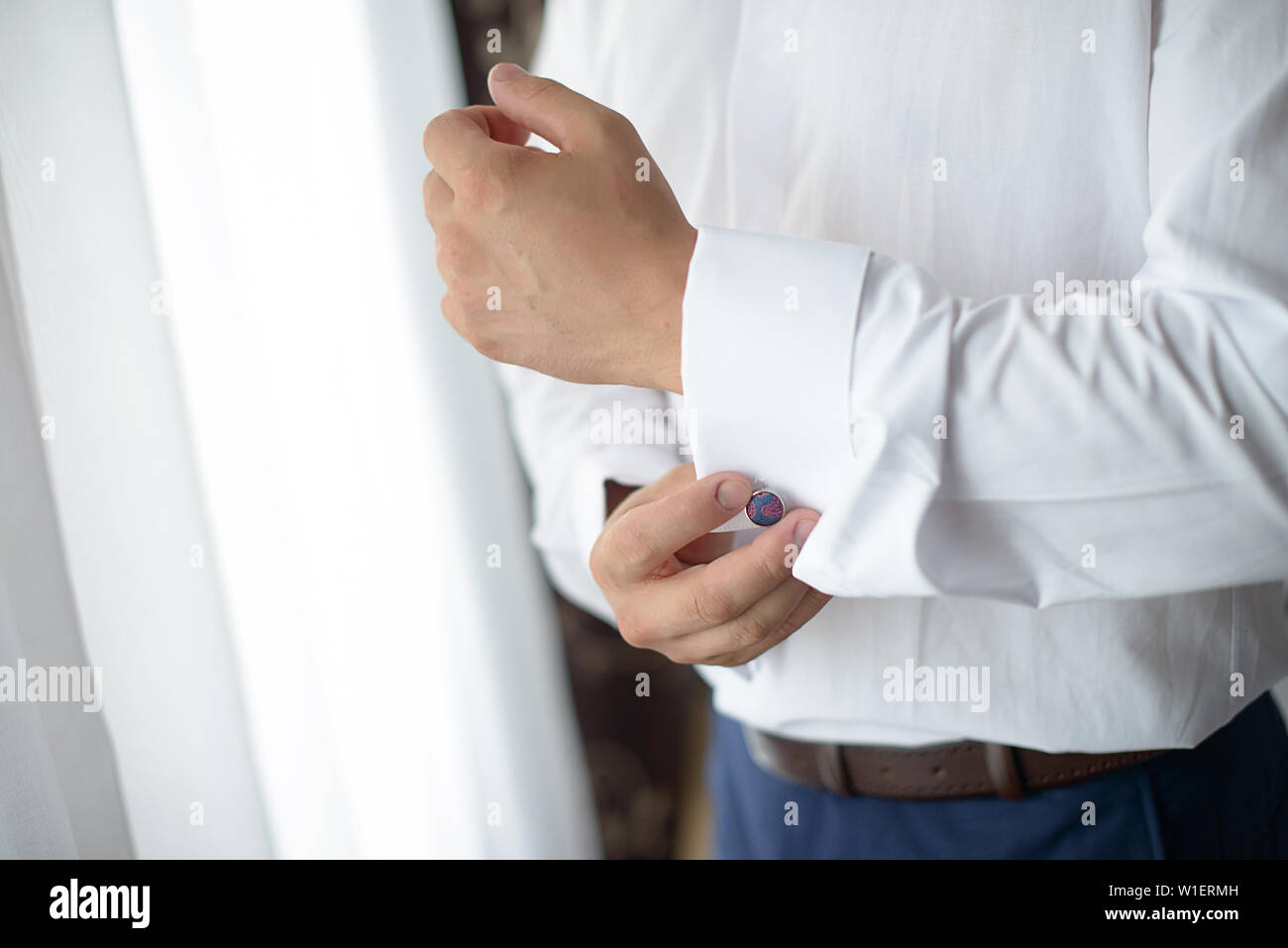 Giovane maschio caucasico vestendosi di fronte a una finestra. Dettagliata orizzontale close-up shot del maschio elegante indossa una camicia bianca che fissa i gemelli Foto Stock