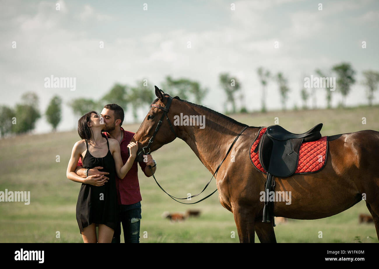 Coppia giovane è abbracciando e baciando in un prato accanto ad un cavallo marrone. Foto Stock