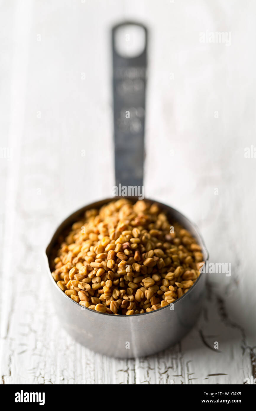 Secchi, materie fenugreek seme nel misurino su bianco cucina rustica sfondo tabella con il fuoco selettivo Foto Stock