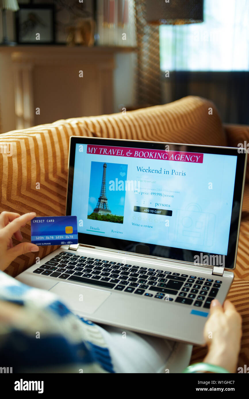 Donna con carta di credito utilizzando online travel hotel prenotazione sito su un computer portatile mentre è seduto sul divano del soggiorno moderno nella soleggiata caldo giorno d'estate. Foto Stock