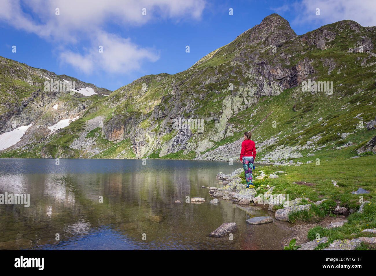 Ragazza con giacca rossa e colorato collant blu con la fotocamera in mano in piedi sul bordo del bellissimo lago di rene sulla montagna Rila Foto Stock