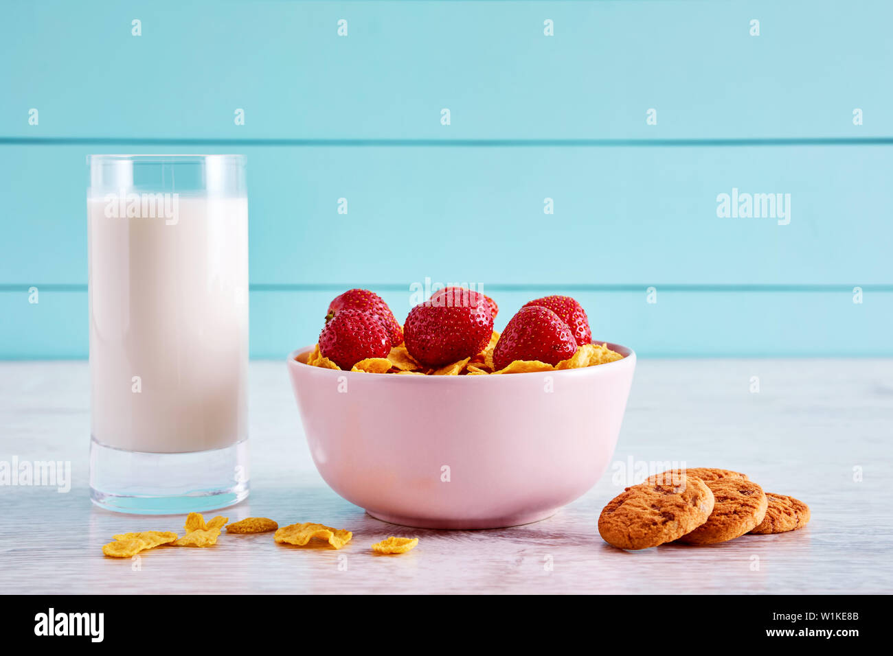 Ciotola di cereali secchi fiocchi di mais con topping di fragole, biscotti al cioccolato e un bicchiere pieno di latte sulla cucina in legno tavolo. Foto Stock