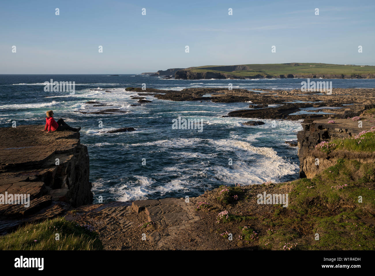 Una donna bionda in una giacca rossa si siede sul bordo della scogliera e gode della vista sul Kilkee e scogliere e l'Oceano Atlantico, Kilkee, County Clare, Foto Stock