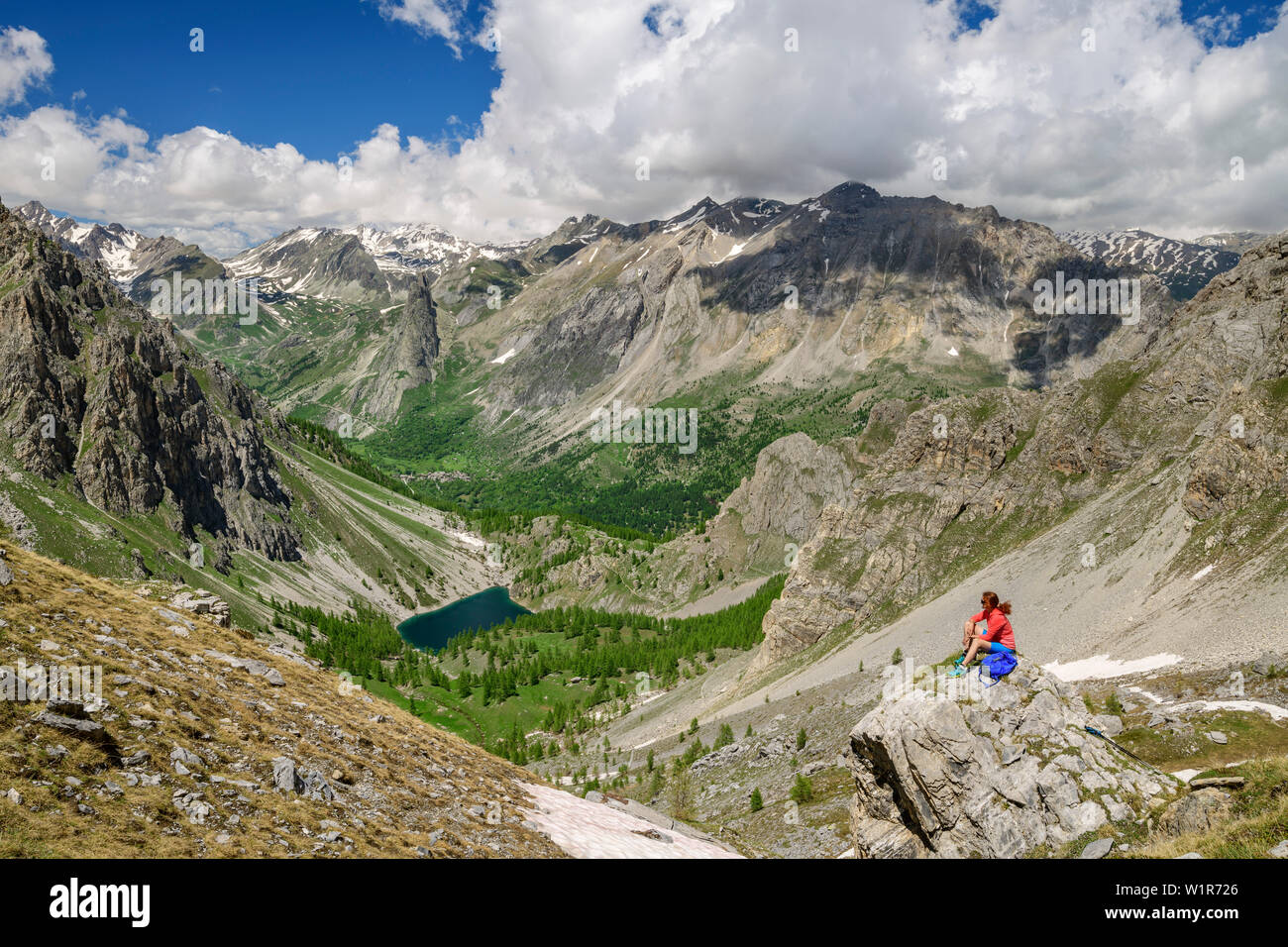Donna seduta escursioni su roccia e guardando verso valle, lago di Montagna ed Alpi Cozie in background, Val Maira, Alpi Cozie, Piemonte, Italia Foto Stock