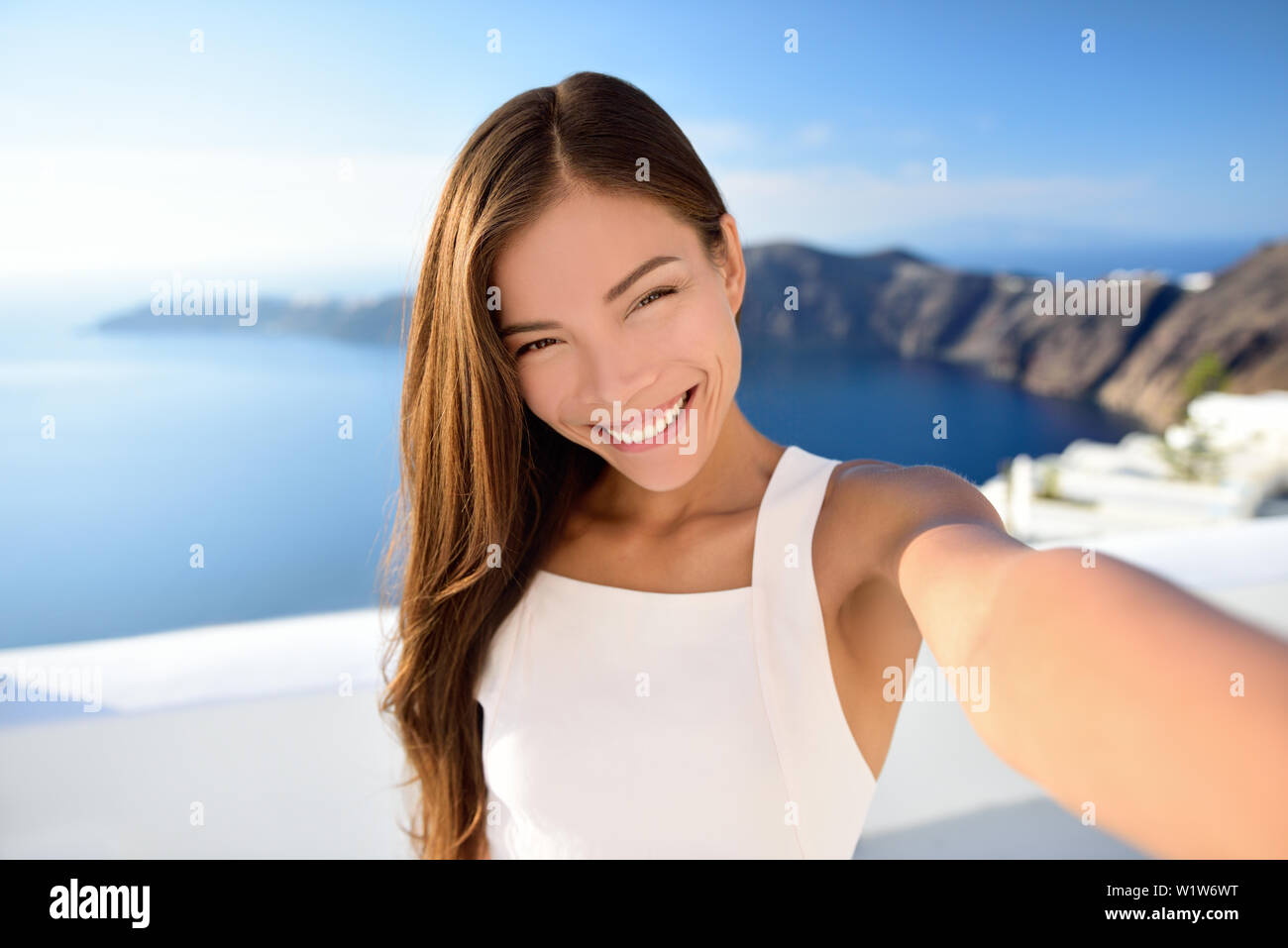 Bellezza asiatica selfie donna su Santorini viaggio estivo. Bella giovane adulto mista cinese di razza caucasica modello femminile di scattare una foto con lo smartphone sorridente alla fotocamera. Dermocosmesi trucco concetto. Foto Stock