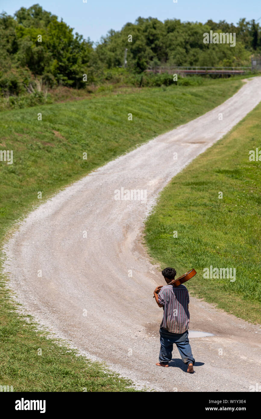 Buras, Louisiana - un afro-americano a piedi nudi uomo che porta una chitarra sulla sua spalla passeggiate lungo una strada di campagna nel sud della Louisiana. Foto Stock