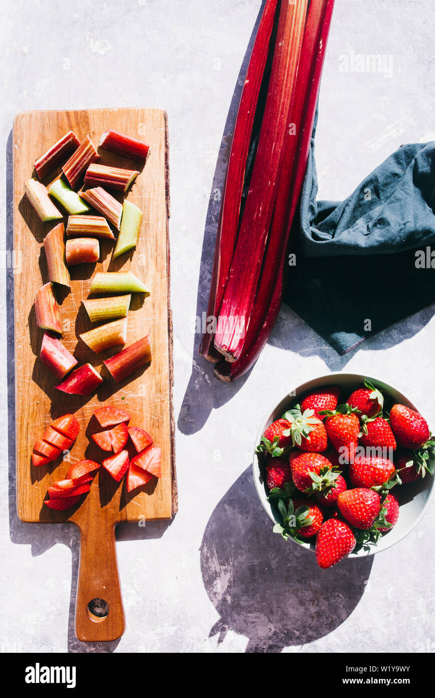 Fresche biologiche il rabarbaro e le fragole tagliate in pezzi su una tavola di legno cutboard su un concreto piano di sfondo scatti, dirigere la luce forte con ricchi sh Foto Stock