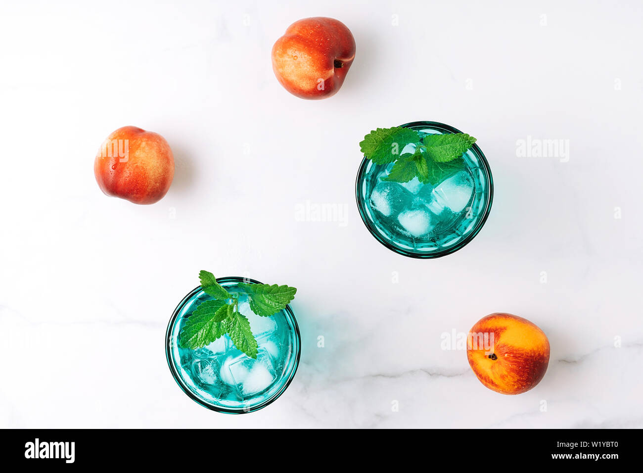 Due belle vintage occhiali turchese con bevanda fredda e cubetti di ghiaccio, decorata con fresco verde menta e tre pesche noci su marmo bianco tabl Foto Stock