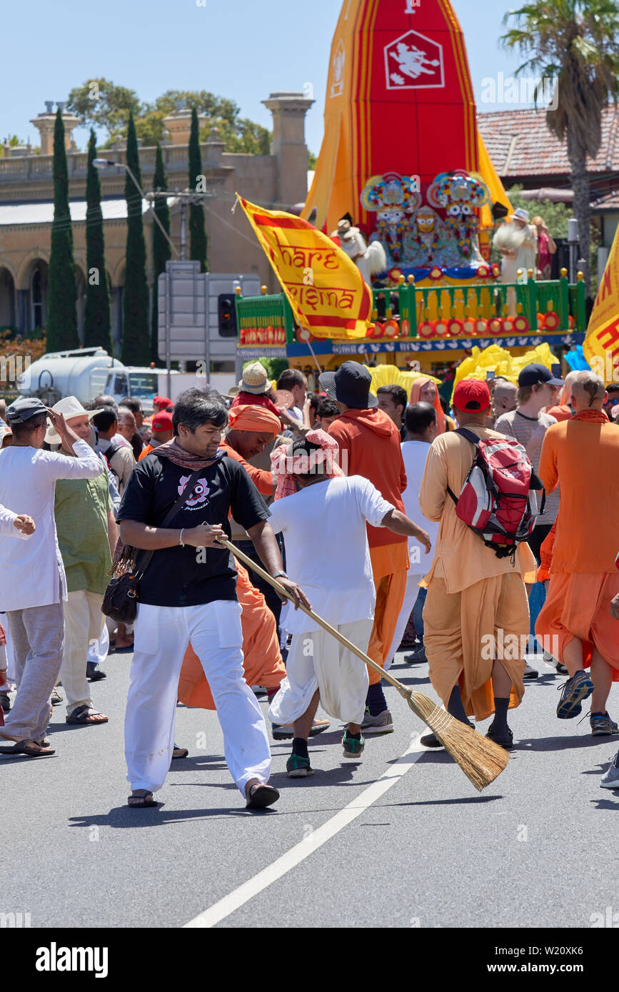 Un uomo spazza la strada utilizzando una scopa di paglia davanti a una lepre Krisha processione durante la St Kilda Festival. Melbourne, Australia - 10 Feb 2019. Foto Stock