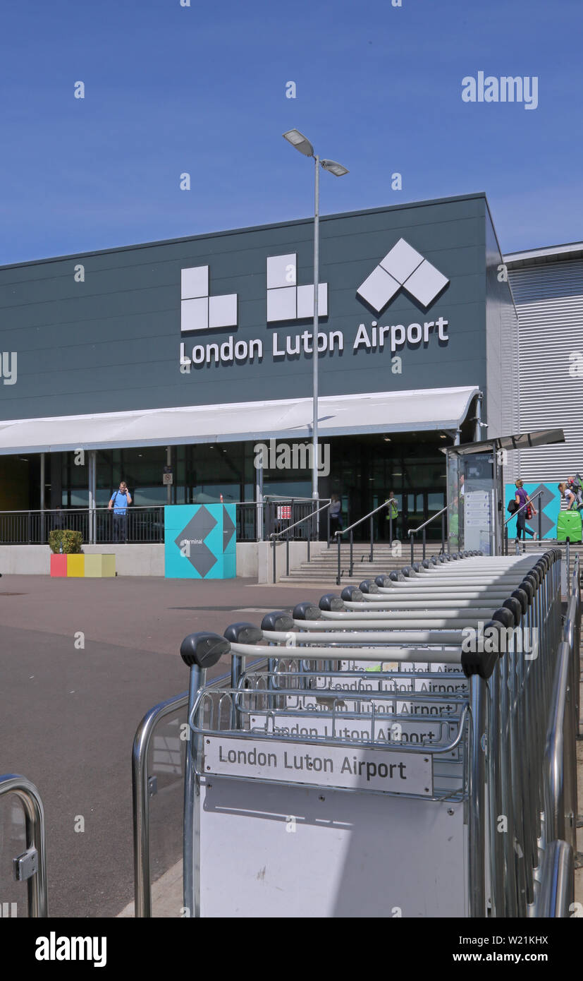 Dall' Aeroporto di Luton a Londra. Carrelli bagagli impilati nella parte anteriore dell'entrata dell'edificio del terminal principale. Mostra il nuovo logo airport (estate 2019) Foto Stock