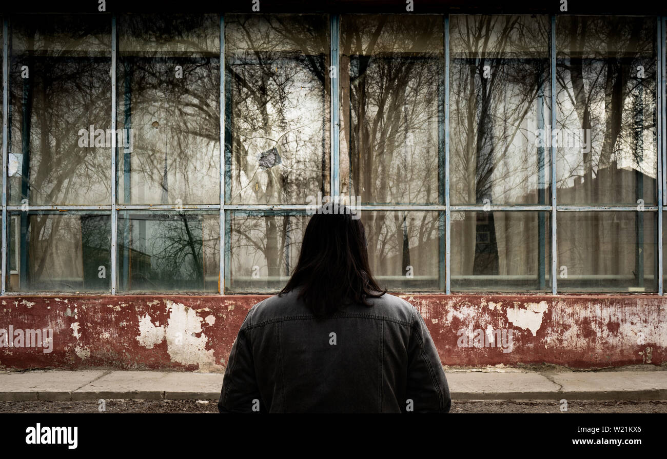 Un uomo con capelli lunghi sta in piedi di fronte ad un edificio in rovina con la sua torna alla fotocamera. Homecoming Foto Stock