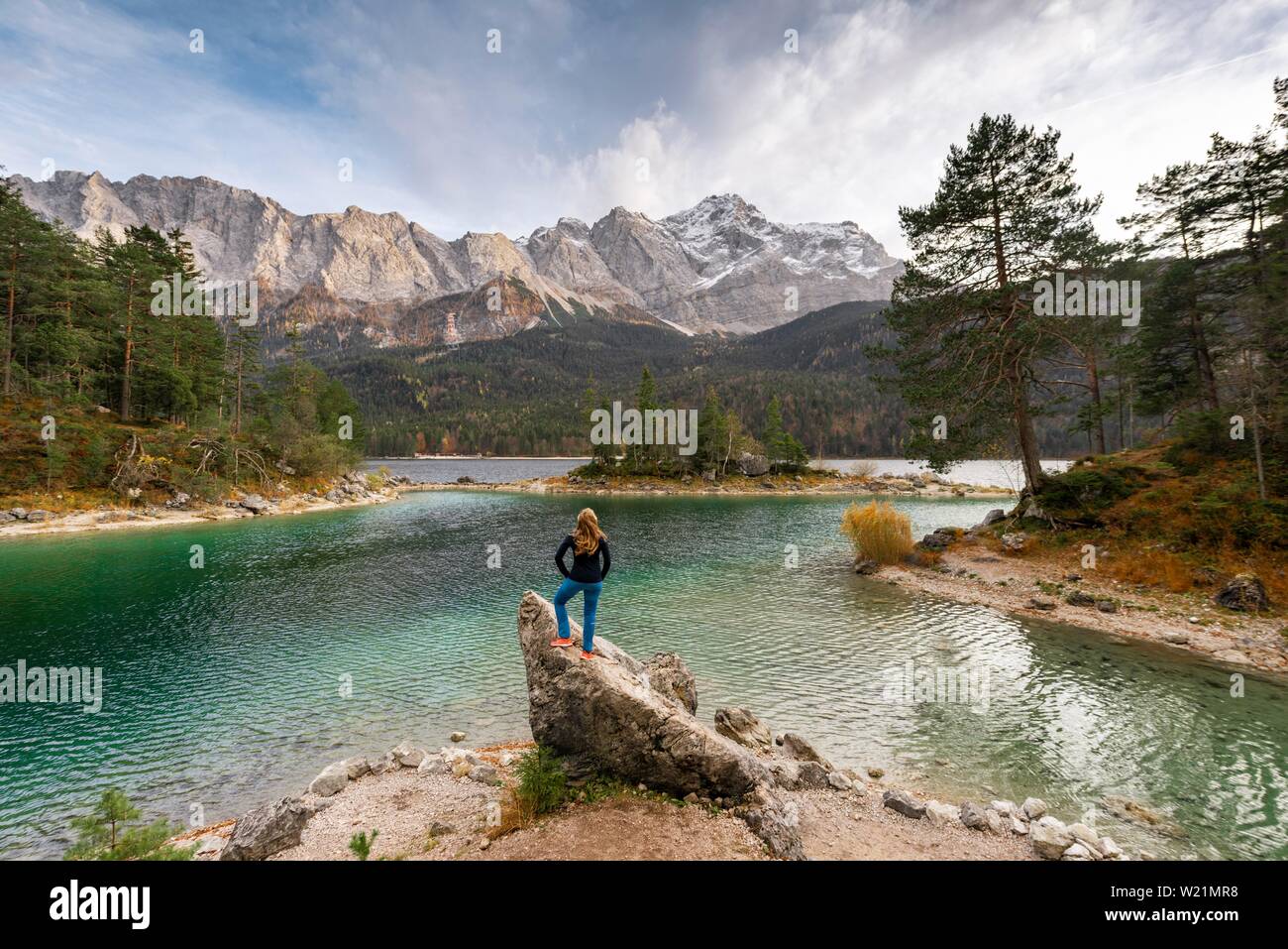 Giovane donna in piedi su una roccia sulla riva, vista in distanza, Eibsee lago di fronte Zugspitzmassiv con Zugspitze, gamma di Wetterstein, vicino Foto Stock