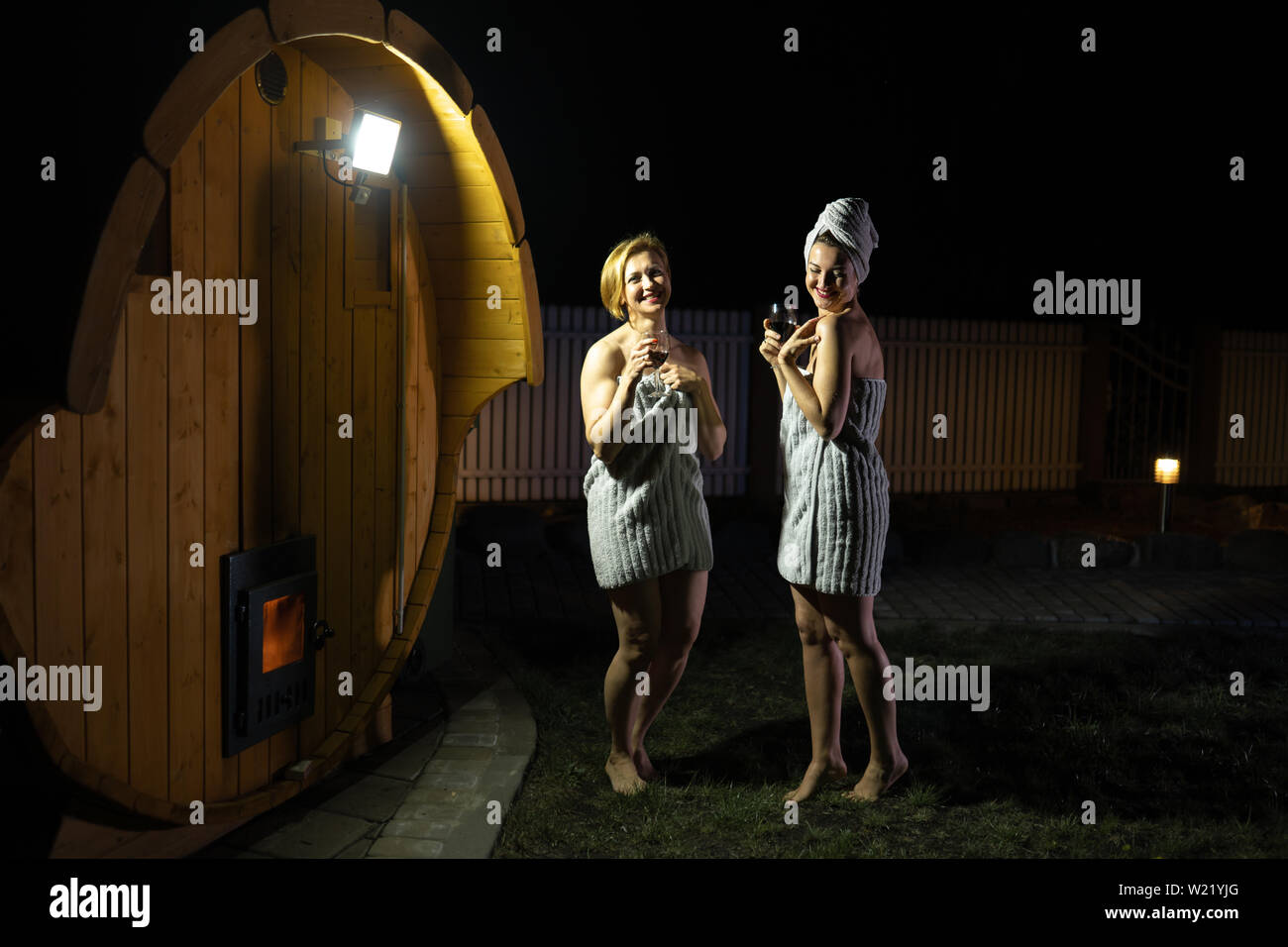 Due amiche donne di bere vino rosso da occhiali fuori nei pressi di un mobile in legno sauna indossando solo asciugamani Foto Stock