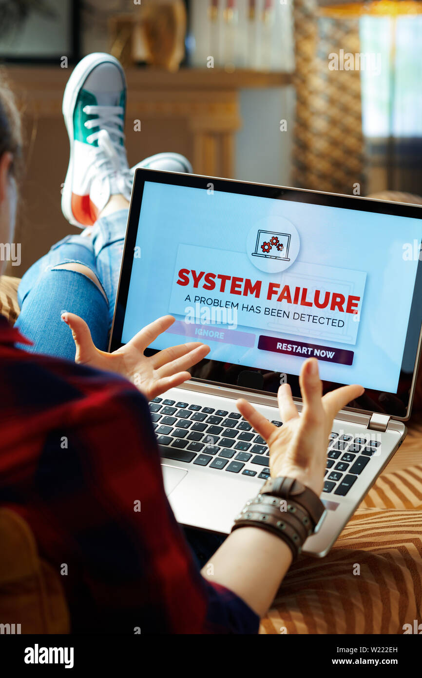 Sulla spalla colpo di infastiditi tanga moderno in rosso a scacchi t-shirt, jeans e scarpe da ginnastica seduti sul divano del soggiorno moderno sperimentando comput Foto Stock