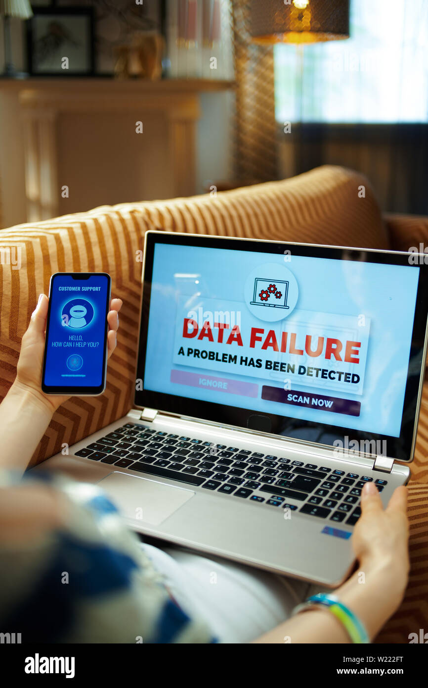 Triste casalinga seduti sul divano a casa moderna in snnny caldo giorno d'estate avente il problema su un computer portatile e di ottenere aiuto dal robot vocale supporto. Foto Stock