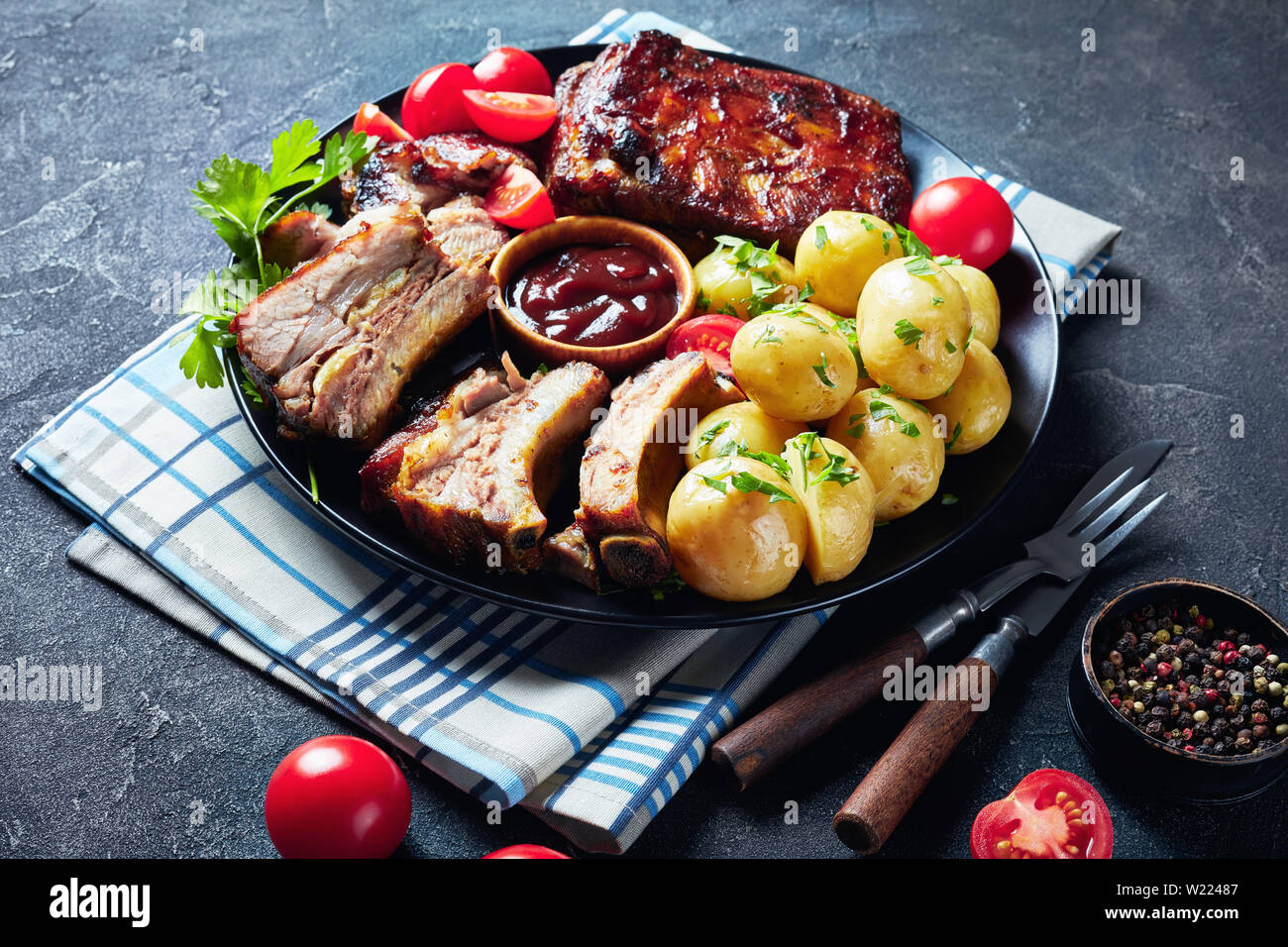 Close-up di grigliate di costolette di maiale condito con salsa barbecue serviti con pomodori e bollito di Hot Potatoes servita su una piastra nera su una tavola di cemento, vie Foto Stock