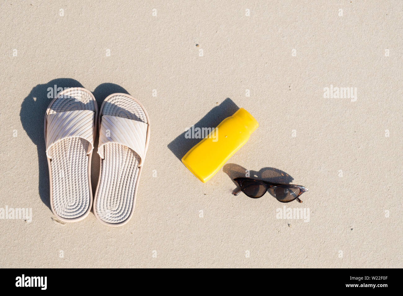 Riposo in spiaggia: pantofole, crema protettiva e occhiali da sole sulla sabbia pulita. Vista dall'alto di accessori per vacanze al mare o vacanze estive Foto Stock