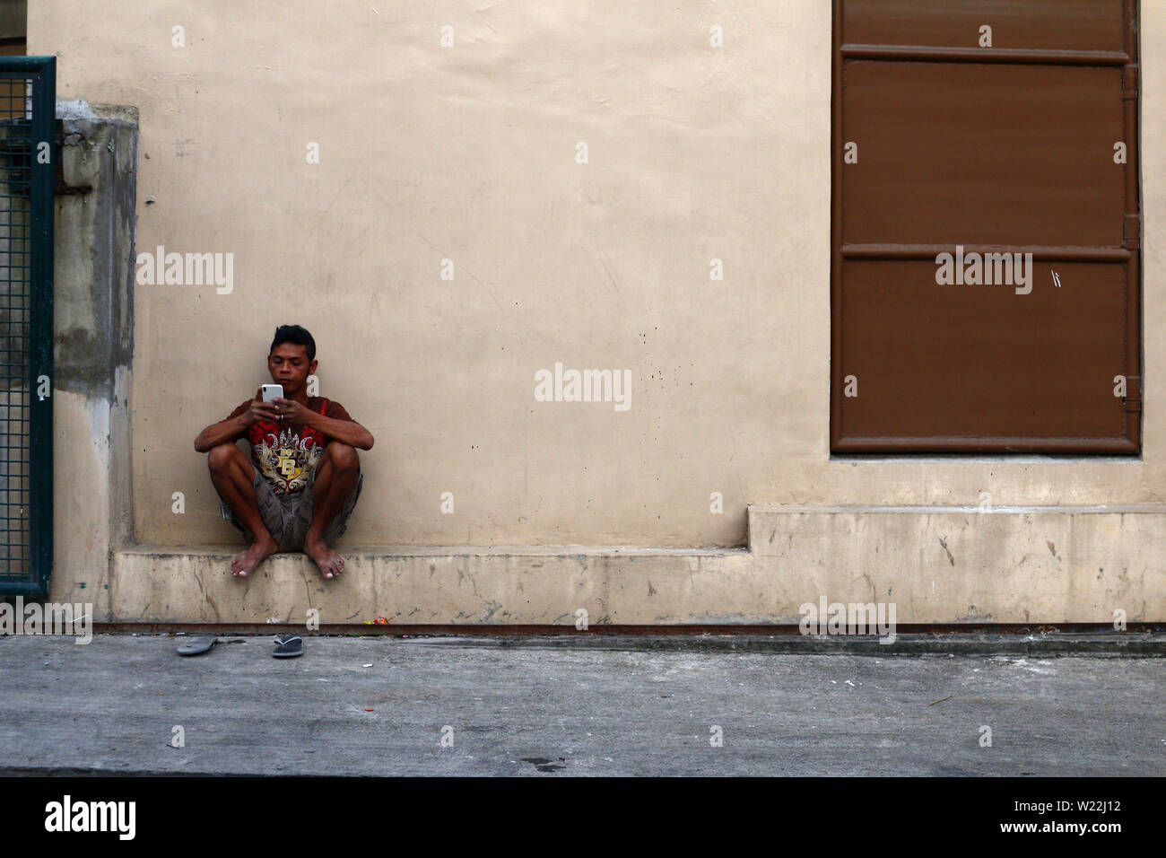 ANTIPOLO City, Filippine - luglio 3, 2019: Un adulto uomo filippino si appoggia al di fuori di un edificio e gioca un gioco sul suo smartphone. Foto Stock
