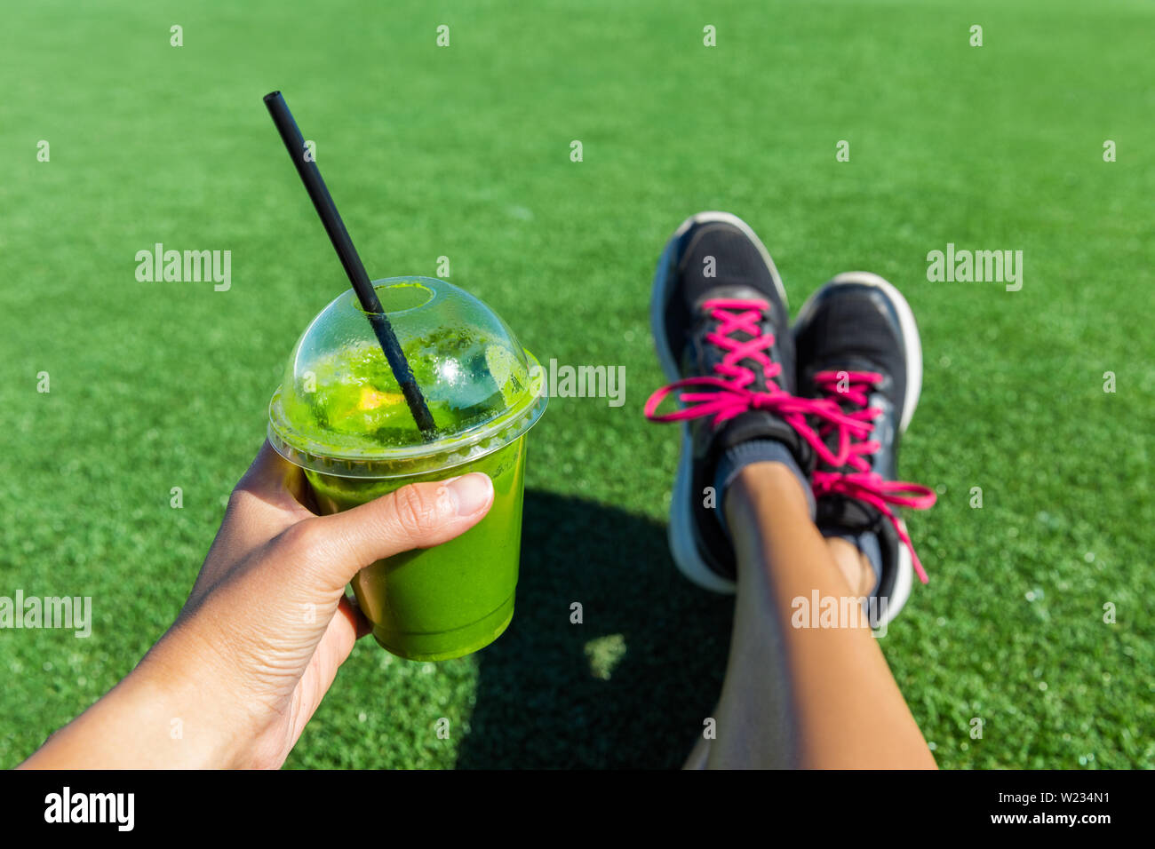 Frullato verde donna bere una tazza di plastica prima colazione da asporto succo di frutta per andare dopo la corsa mattutina nel parco d'estate. Uno stile di vita sano persona sportiva pov di mano azienda bere con scarpe da corsa piedi selfie. Foto Stock