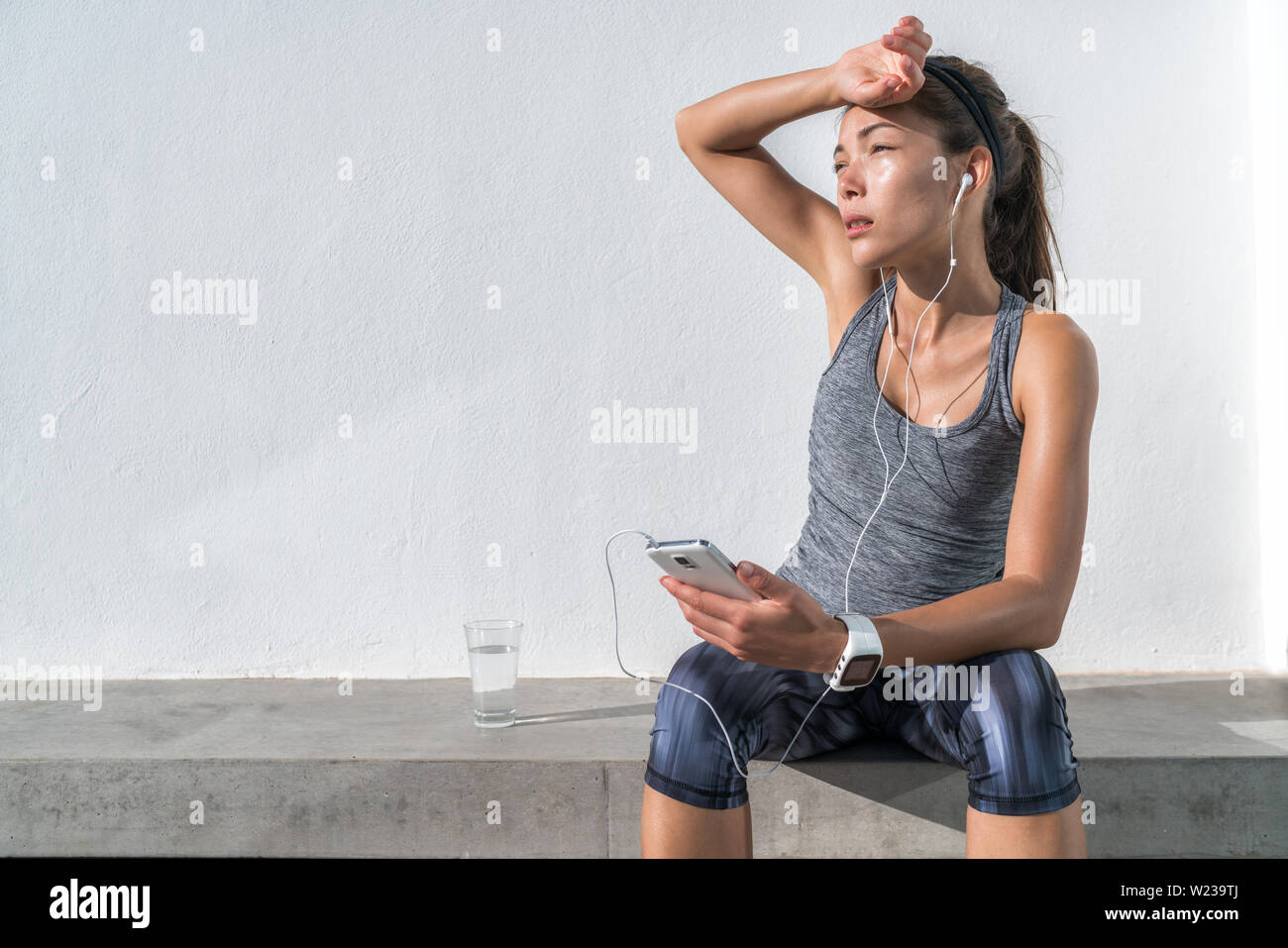 Stanco donna fitness sudorazione prendendo una pausa per ascoltare musica sul telefono dopo il difficile percorso di formazione. Esaurito Asian runner disidratato sensazione di stanchezza e disidratazione dal lavoro in palestra. Foto Stock