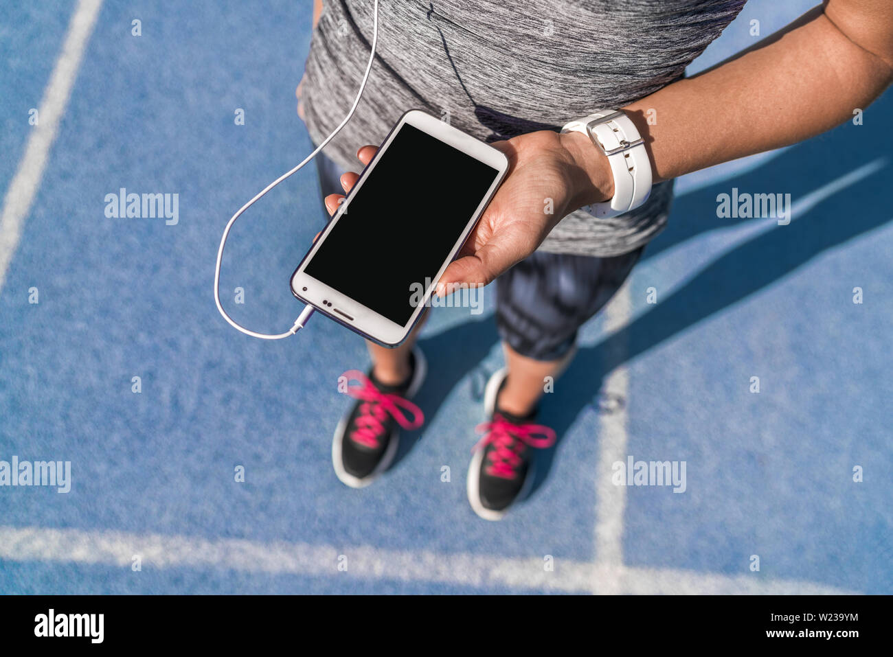 Runner ragazza tenendo lo smartphone utilizzando il touchscreen per la scelta della musica o i messaggi di testo SMS sulla prima app in esecuzione su pista. Atleta femminile donna piedi e gamba closeup con smartwatch e mano toccando il display. Foto Stock