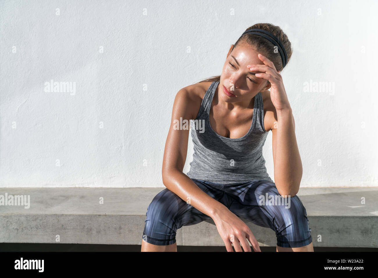 Stanco di fitness donna in esecuzione sudorazione prendendo una pausa di allenamento cardio training difficile. Esaurito runner seduta con mal di testa sensazione di stanchezza e disidratazione dal lavoro in palestra a cielo aperto. Foto Stock