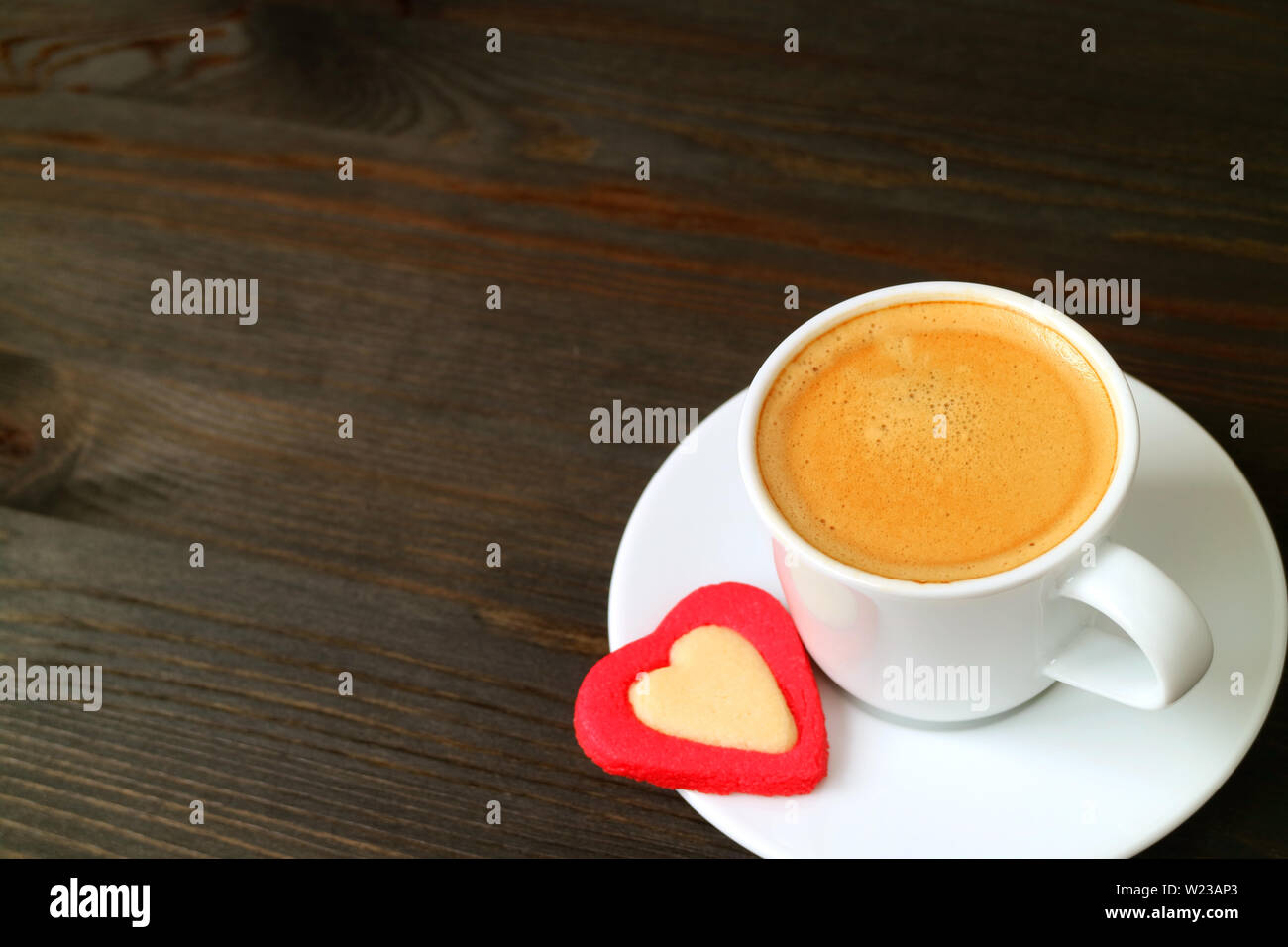 Una tazza calda di caffè espresso con un cuore cookie sagomato in legno marrone scuro e tavolo con spazio di copia Foto Stock