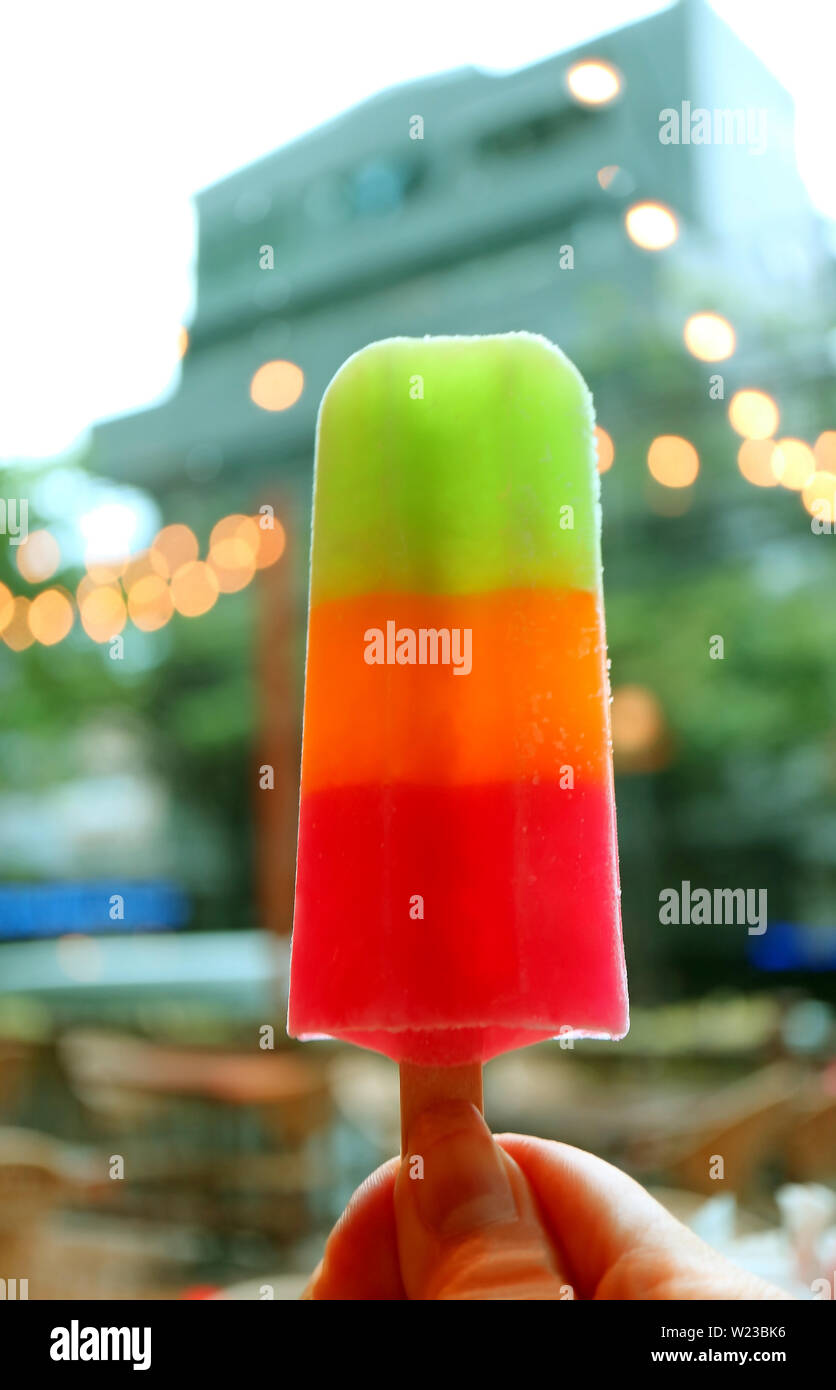 Donna di mano che tiene una frutta mix ice bar contro sfocata edificio con luci bokeh di fondo Foto Stock