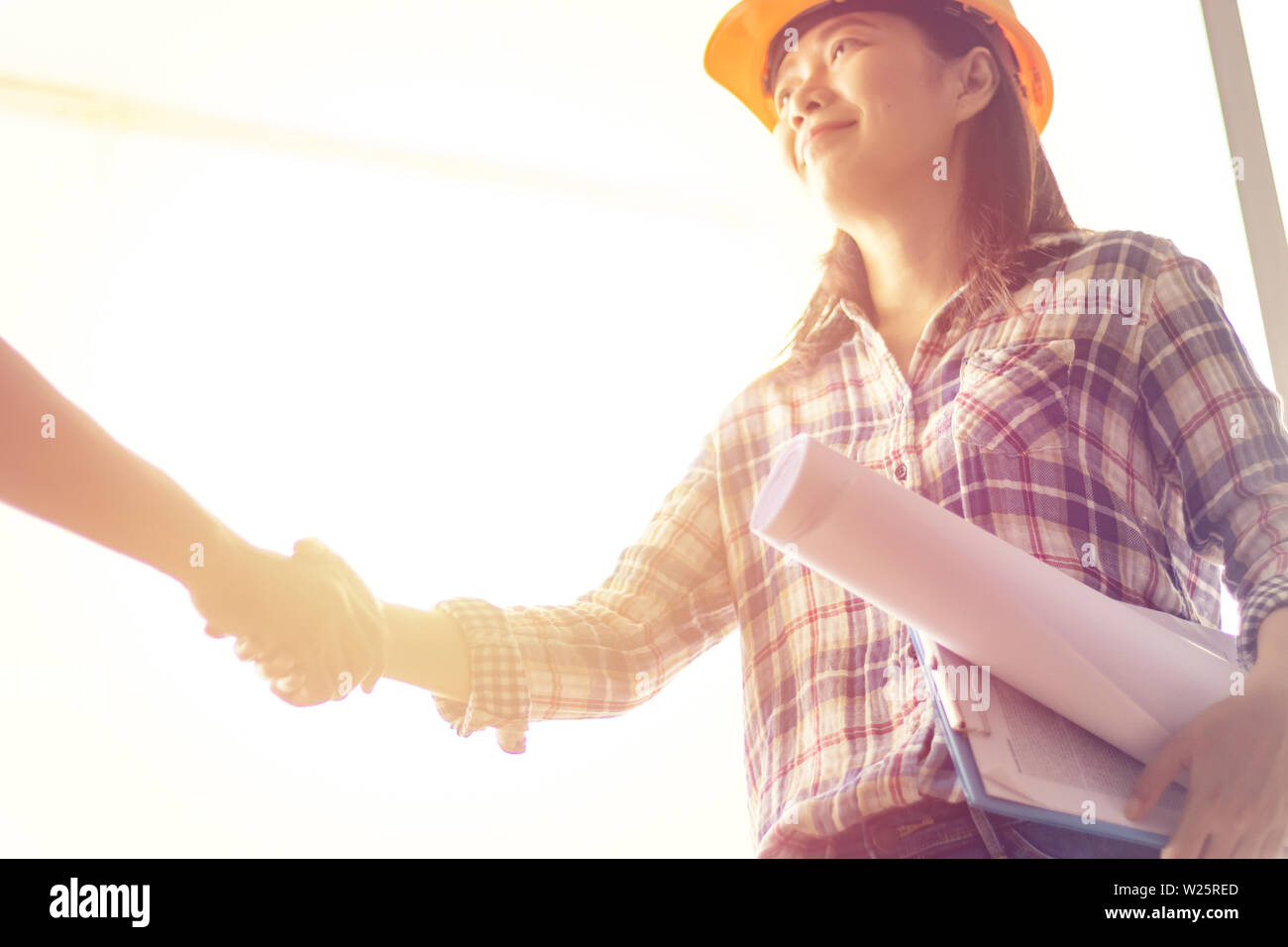 Felice donna asiatica ingegnere come lavoratori edili con blueprint e contratto in mano con protezione Casco giallo si stringono la mano durante il lavoro Foto Stock