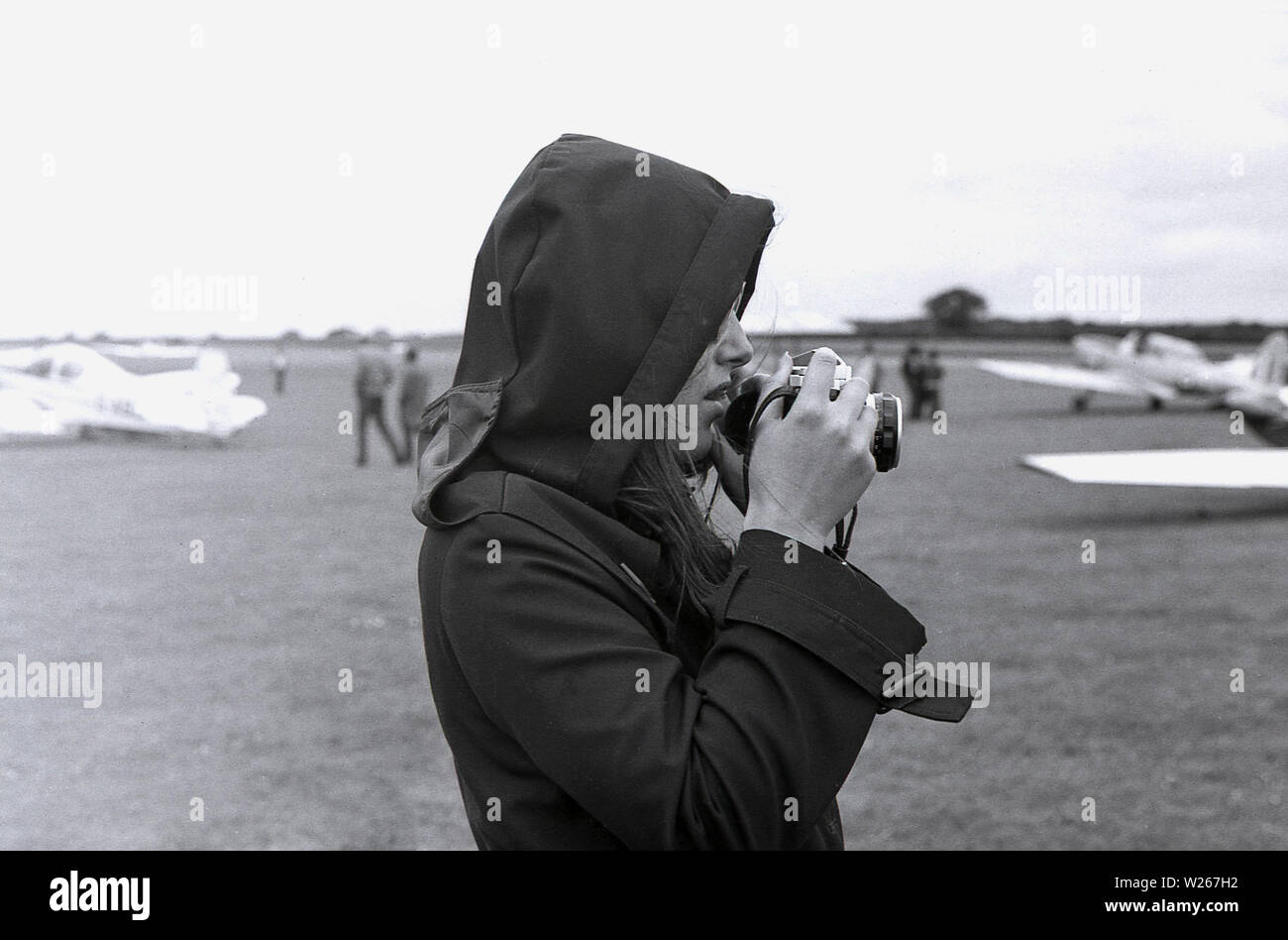 Anni Settanta, storico di una giovane donna che indossa una giacca a vento con il suo cofano fino oltre i suoi capelli, tenendo una cinepresa, scattare fotografie presso un'esibizione aerea, Old Warden Aerodrome, Biggleswade, Inghilterra, Regno Unito. Foto Stock